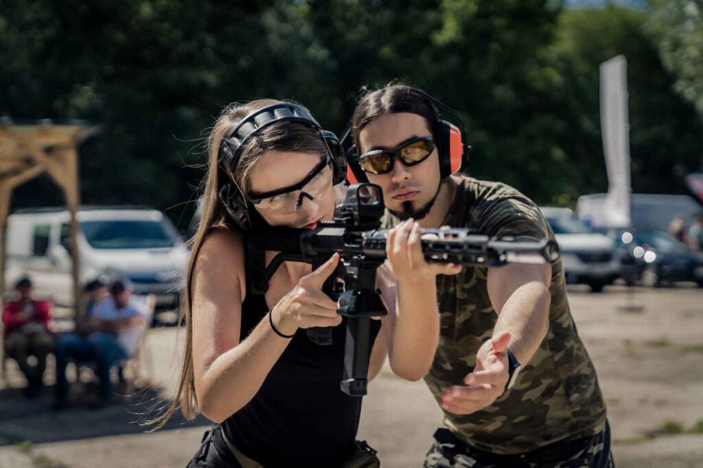 Shooting range concept. Private shooting lessons with an instructor. Medium outdoor shot of a powerful long-haired woman wearing protective equipment at aiming at the target with black riffle with collimator. High quality photo